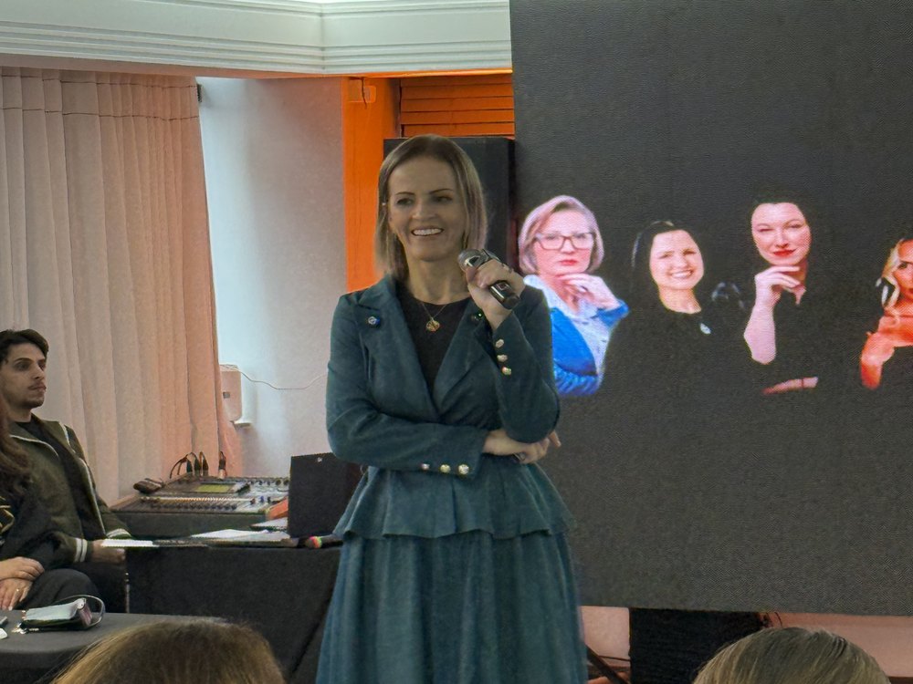 Luciene Jokoski falando ao microfone diante de uma plateia, durante palestra